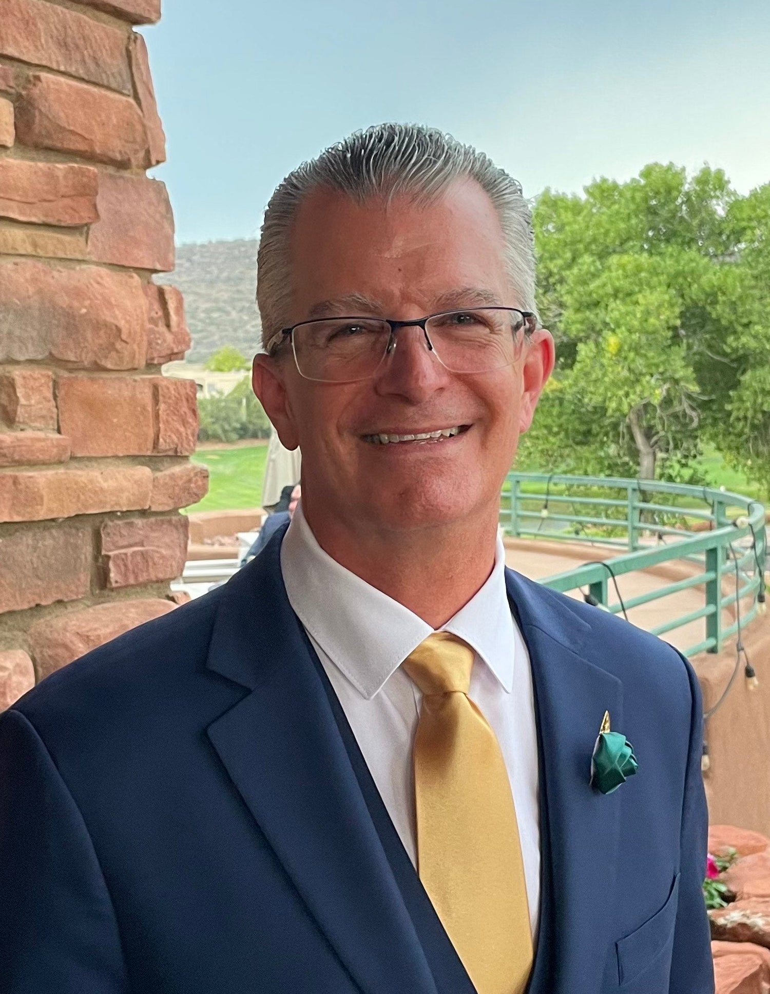 Man in a suit standing next to a brick wall with trees in the background
