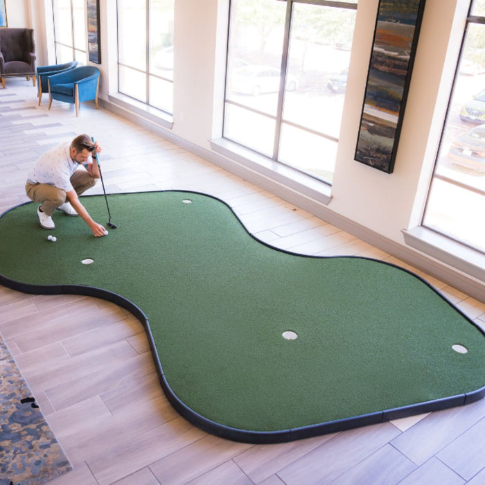 Person playing indoor putting green in a room with large windows.